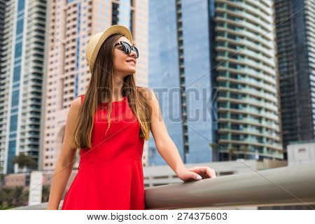 Portrait Of Young Woman Wearing In Red Dress And Sunglasses, Straw Hat In Front Of Skycrapers In Mod