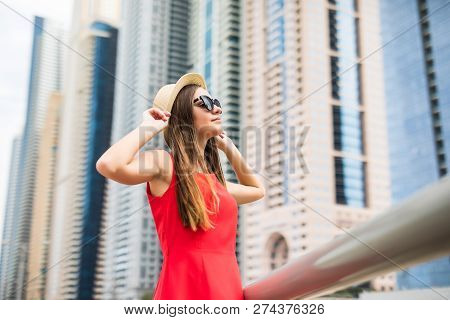 Young Pretty Woman Holding Straw Hat Wearing In Red Dress And Sunglasses In Front Of Skycrapers.
