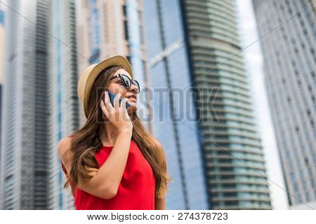Portrait Of Young Smile Woman In Red Dress, Sunglasses And Summer Hat Talking On The Phone On Downto
