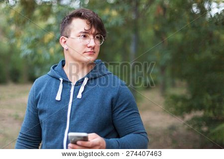 Attractive Young Caucasian Man With Dark Curly Hair, Golden Round Glasses And Earring Holds The Mobi