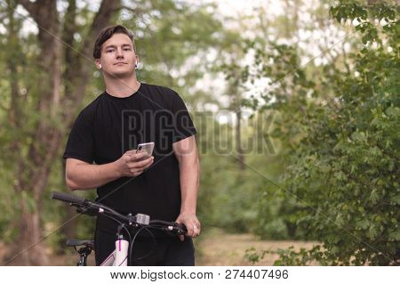 Attractive Young Caucasian Man Holds Mobile Phone And Bicycle. White Earphones, Earring, Dark Casual