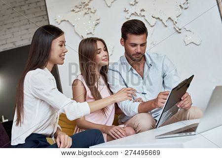 Young Lovely Couple Sitting With Manager At The Travel Agency Office And Reviewing Contract For A Su