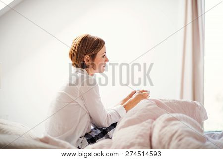 A Young Woman With Night Shirt Sitting Indoors On Bed In The Morning, Holding Cup.