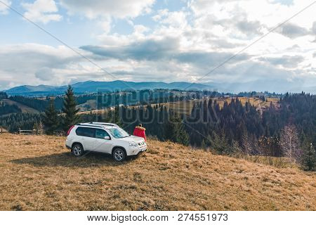 Man In Red Coat Near White Suv Car At Hill With Beautiful Mountains View. Road Trip.