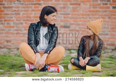 Shot Of Pleased Realxed Two Sisters Pose Crossed Legs, Sit On Lawn, Pose Against Brick Wall Backgrou