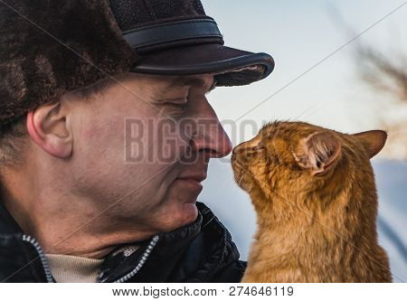 A Middle-aged Man In A Brown Hat And A Red Cat Look At Each Other In Winter