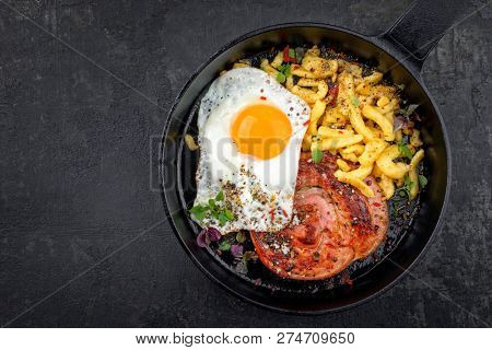 Traditional fried Allgauer veal roast roll sausage slices with fried egg and spaetzle in a cast-iron skillet with spices as closeup on a black board