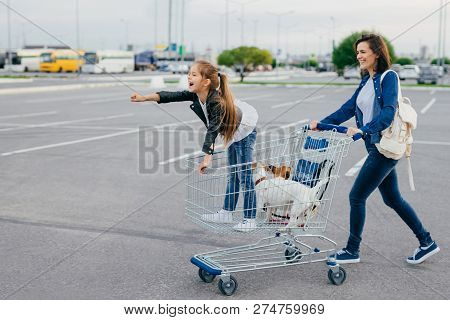 Photo Of Happy Small Girl With Her Mother And Pets Stands In Cart, Make Shopping During Weekend, Enj