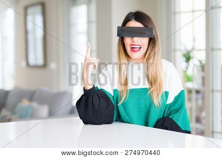 Young beautiful woman blindfold eyes with black censor band at home pointing finger up with successful idea. Exited and happy. Number one.