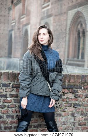 Attractive young woman wearing gray dress and hakki jacket with long fair hair posing outdoor over ancient Castello Sforzesco, Milan, Italy. Beautiful caucasian model in old italian city.