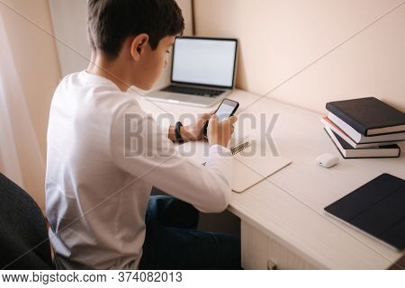 Schoolboy Study At Desk In His Room. Boy Use Laptop And Smartphone To Writing In Notebook. Books And