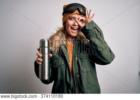 Young beautiful skier woman with blue eyes wearing ski goggles drinking thermo with coffee with happy face smiling doing ok sign with hand on eye looking through fingers