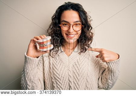 Young beautiful woman with curly hair holding plastic teeth with dental baces with surprise face pointing finger to himself