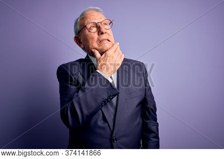 Grey haired senior business man wearing glasses and elegant suit and tie over purple background Thinking worried about a question, concerned and nervous with hand on chin