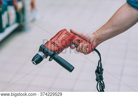 A Man Holds An Electric Drill In His Hand For Drilling. A Buyer In A Hardware Store Selects A Produc