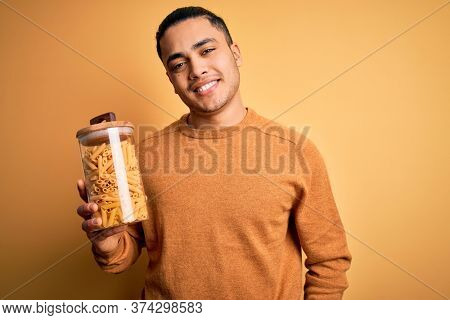 Young brazilian man holding jar with Italian dry pasta macaroni over isolated yellow background with a happy face standing and smiling with a confident smile showing teeth