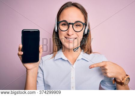 Beautiful call center agent woman working using headset holding smartphone holding screen with surprise face pointing finger to himself