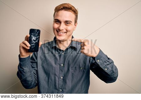 Young handsome redhead man holding broken smartphone showing cracked screen pointing finger to one self smiling happy and proud
