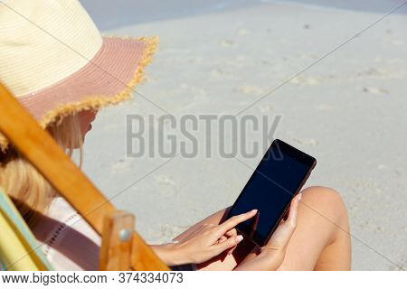 Attractive blonde Caucasian woman enjoying time at the beach on a sunny day, sitting on deck chair, using a tablet computer, with blue sky and sea in the background. Summer tropical beach vacation.