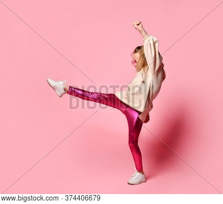 Side View Of Little Girl In Pink Hoodie, Sparkly Leggings And Glasses Standing On One Leg While Exte