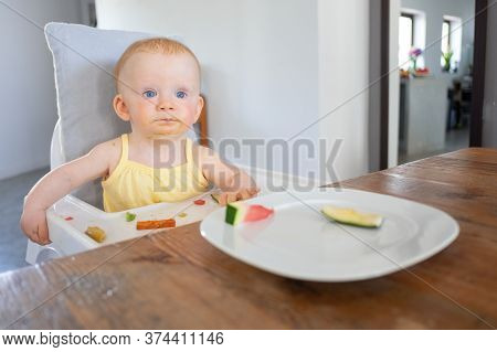 Adorable Baby Girl Sitting In Highchair With Food Messy On Tray And Face, Trying To Eat Pieces Of Fr