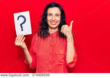 Young beautiful hispanic woman holding question mark smiling happy and positive, thumb up doing excellent and approval sign 
