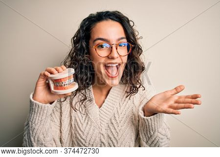 Young beautiful woman with curly hair holding plastic teeth with dental baces very happy and excited, winner expression celebrating victory screaming with big smile and raised hands