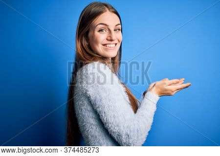 Young beautiful redhead woman wearing casual sweater over isolated blue background pointing aside with hands open palms showing copy space, presenting advertisement smiling excited happy