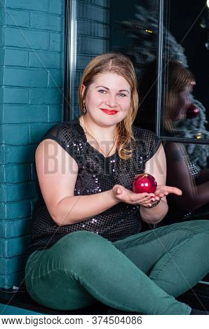 Happy, Smiling Woman With A Christmas Ball, Decoration In Her Hands Sits In A Room On The Windowsill