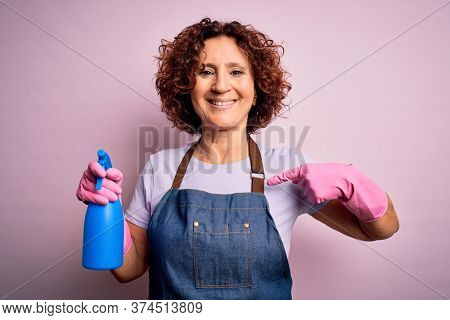 Middle age curly hair woman cleaning doing housework wearing apron and gloves using spayer with surprise face pointing finger to himself