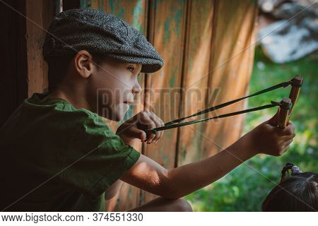 Child Boy In A Cap Takes Aim With A Slingshot To Shoot At A Target. Play Around As A Child In The Vi