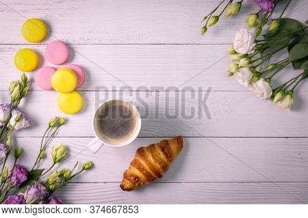 Macarons And Croissant With Flowers And Cup Of Coffee On White Wooden Background With Copy Space. To