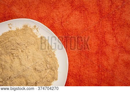 maca root powder on white ceramic plate against red textured paper, superfood concept