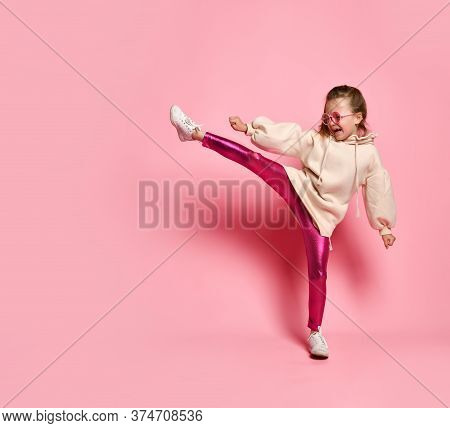 Side View Of Little Girl In Pink Hoodie, Sparkly Leggings And Glasses Standing On One Leg While Exte