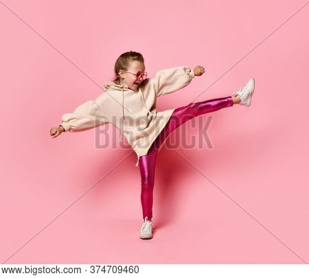Side View Of Little Girl In Pink Hoodie, Sparkly Leggings And Glasses Standing On One Leg While Exte