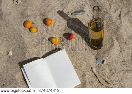 A Bottle Of White Delicious Wine, Five Juicy Apricots And A Book With Blank Sheets Of Paper On Sand 
