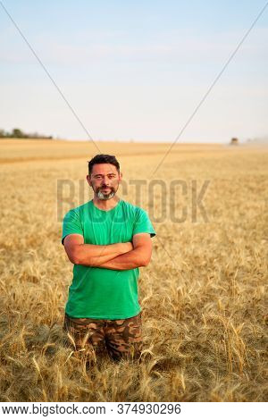 Happy Farmer Proudly Standing In Wheat Field With Arms Crossed On Chest. Agronomist Wearing Corporat