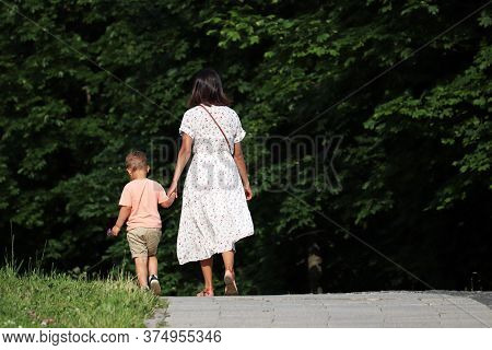 Mother And Kid Are Walking Down The Path Holding Hands. Young Woman In Dress Lead Little Boy In A Su