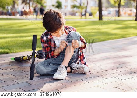 A Little Boy 8 Years Riding Scooter And Fell. Sitting On Asphalt, Holding Hands On His Injured Leg A
