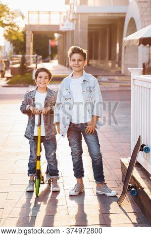 two attractive teenagers of Caucasian nationality 7 and 9 years old standing on sunny evening street in the city. Laughing, pointing fingers forward, holding scooter and skateboard