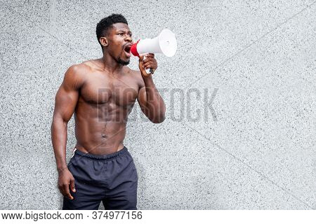 African American Man With An Athletic Body Stands Outdoors Against A Wall And Shouts Into A Megaphon