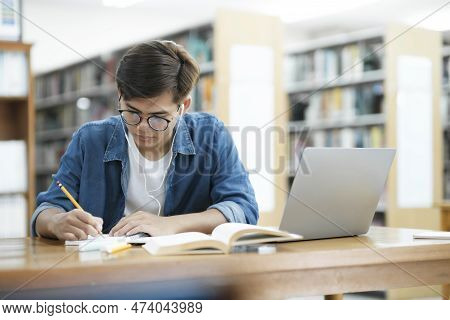 Young Male College Student Wearing Eyeglasses And In Casual Cloths Sitting At Desk Studying, Reading