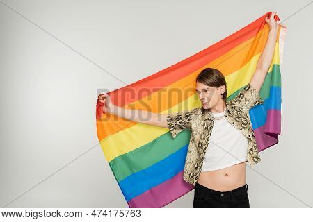 Cheerful And Stylish Pangender Person Standing With Rainbow Flag And Looking Away Isolated On Grey.