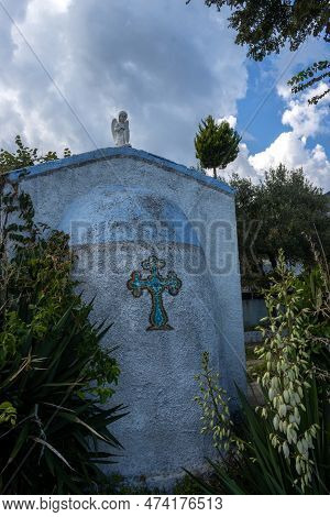 Blue Chapel With A Cross, In A Garden At The Coast. White Angel On The Roof. Blooming Yucca. Blue Sk