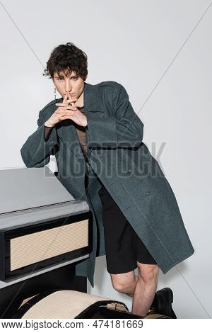 Thoughtful Pangender Person In Coat Posing With Clenched Hands Near Model Of Photo Camera On Grey Ba