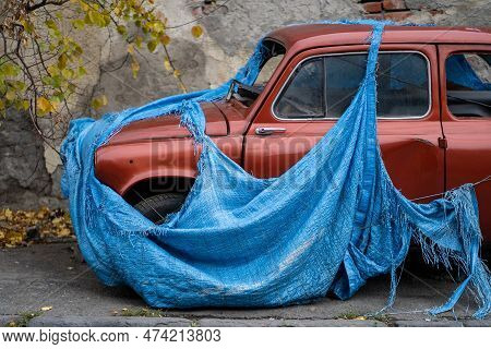 Retro Old Abandoned Red Car With Broken Glass Covered With Torn Blue Awning To Prevent Metal Corrosi