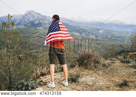 Young Man Standing On Rock Cliff With The Us Flag On His Back While Looking At Mountains In Backgrou