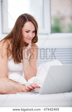 美丽快乐的年轻女人在床上和在家中使用笔记本电脑 sittting