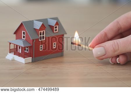 Hand Of Woman Holding Burning Match Near Two-story Paper House Standing On Wooden Table. Bright Fire