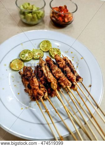 A Plate Of Chicken Satay Served With Lime, A Glass Of Pickles And Sambal Matah In The Room, Their Vi